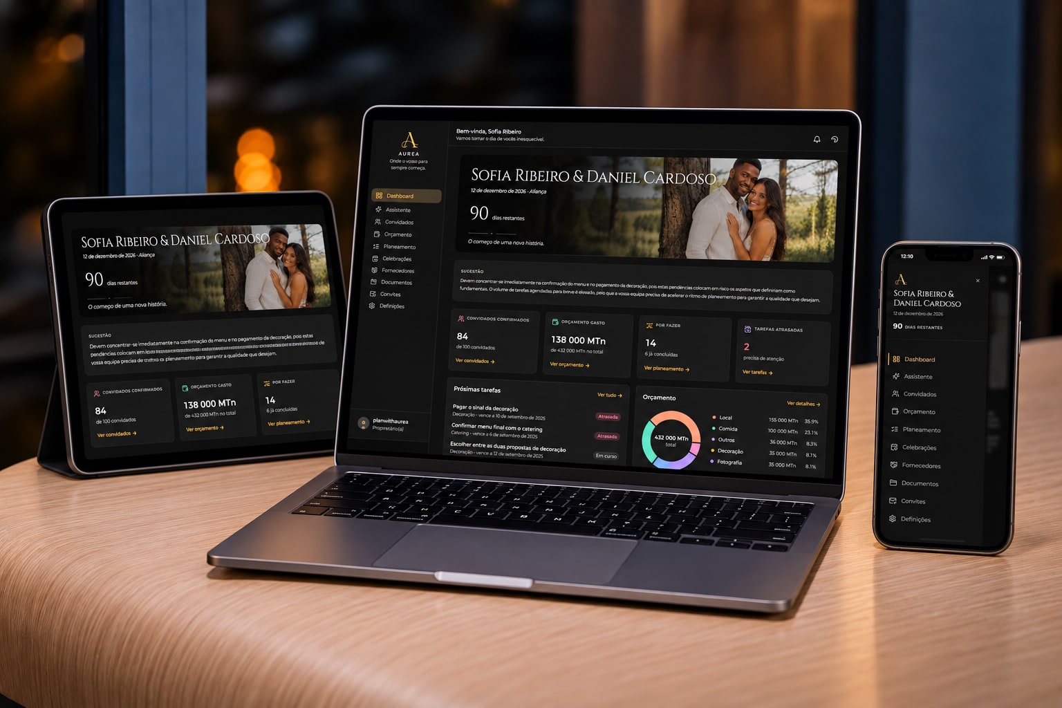 A Aurea aberta em três aparelhos sobre uma mesa: um MacBook ao centro com o painel do casamento, um iPad à esquerda e um iPhone à direita.